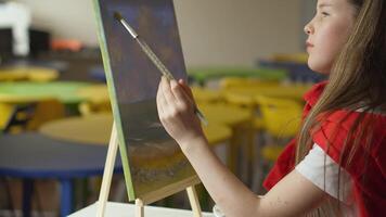 A focused girl is painting a landscape with a brush on a canvas propped on a wooden easel. The classroom setting includes colorful tables, highlighting a creative learning environment. video