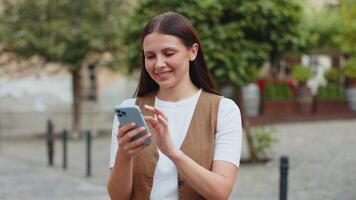 Smiling young woman using smartphone browsing internet, finishing work, looking at camera outdoors video