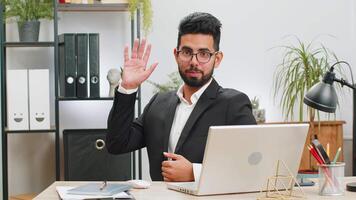 Businessman working on laptop smiling friendly at camera and waving hands gesturing hello at office video