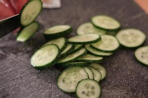 Fresh cucumber slices being cut on a wooden cutting board in a kitchen setting during meal preparation photo
