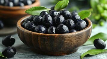 Fresh black olives in wooden bowl with green leaves on gray surface for healthy cooking and snacking ideas photo