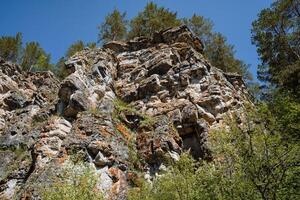A majestic rocky cliff adorned with vibrant trees scattered across its surface, complemented by a breathtaking blue sky gleaming in the background, creating a picturesque natural scene photo