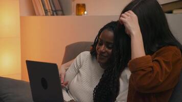 Multiracial lesbian couple sitting on sofa in cozy evening setting, emotionally reacting and discussing surprising content on laptop, sharing a warm and intimate moment together. video