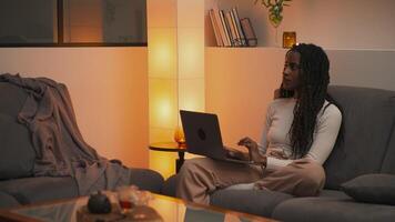 Young African American woman with dreadlocks sitting on sofa at home in the evening, calmly working on laptop with thoughtful expression and gentle smile in a cozy, relaxed setting. video