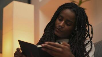 Close-up portrait of young African American woman with dreadlocks reading a book and turning pages in soft evening light, immersed in a calm and cozy atmosphere. video
