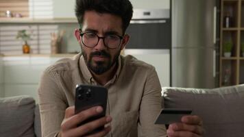Arabian Indian man on sofa confused upset business client at home kitchen problem online payment credit limit use mobile phone app and bank card failure mistake error trouble not enough money balance video