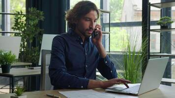 Arabian man hispanic businessman manager employer talk mobile phone search information on laptop check documents on desktop consulting client remote business consultation multitasking work in office video