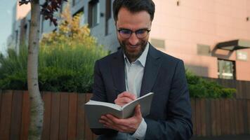 Caucasian business man businessman outside thinking writing notes in notebook university teacher professor in city hold paper notepad smile come up with idea creative thought prepare for exam studying video