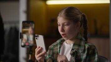 Joyful Caucasian little kid girl child influencer blogger schoolgirl record blog on camera tripod review new smartphone mobile phone device opinion showing thumb up approve like gesture at home video
