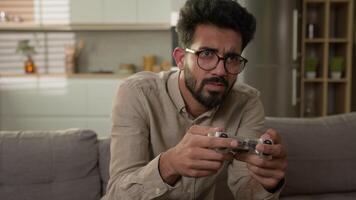 Arabian Indian man male guy gaming addiction play online console videogame on TV playing virtual sport with controller on couch sofa at home kitchen annoyed gamer disappointed about losing game over video