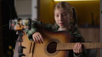 Creative Caucasian little kid girl child influencer blogger schoolgirl explain teaching how to playing guitar singing song record online tutorial blog mobile phone talking camera tripod at home video