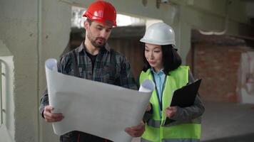 Multiethnic builders in uniform discussing blueprint inspection business engineers Caucasian man Korean woman hardhat discuss plan construction building project two workers partners technicians indoor video