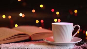 On a dark table covered with artificial snow, there is a white cup and an old open book. video