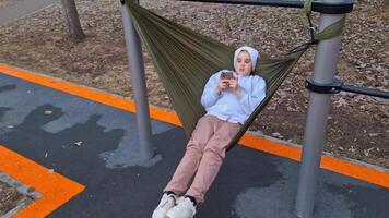Girl relaxing with smartphone on outdoor hammock in urban park video