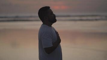 Brown Beard Asian man practices breath control at sunset on the beach. A quiet moment of healing, mindfulness, emotional grounding, and inner peace. Dark video