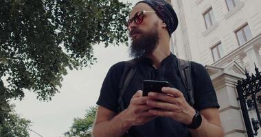 A young man sporting a beard is using his smartphone outdoors while enjoying the daylight video