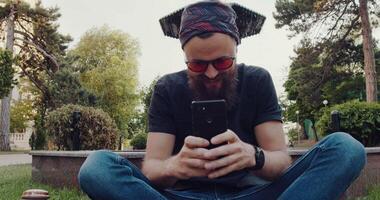 A bearded man sits on the grass, focused on his cell phone video