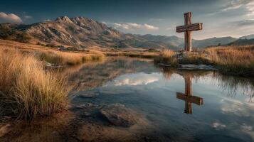 Wooden cross stands by the tranquil pond reflecting the majestic mountains at sunset in a serene landscape photo