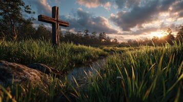 Wooden cross stands amidst tall grass and rocks near a peaceful stream during sunset in a serene natural landscape photo