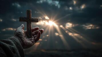 Hand holding wooden cross against a dramatic sky with sunlight breaking through clouds at dawn photo