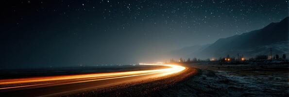 Nighttime coastal road with illuminated car trails under a starry sky near mountains photo