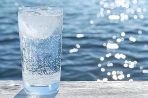 Refreshing glass of sparkling water with ice, ocean backdrop photo
