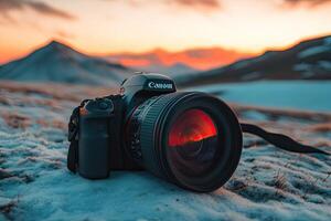 A camera rests on snowy ground, facing sunset over mountains photo