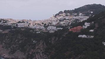 Aerial view of Capri, Italy, showing rugged cliffs, a hillside town with whitewashed buildings, and the sea. Smooth motion captures the overcast scene. video