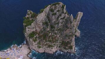 Sweeping aerial view of Capri, Italy, showcasing cliffs, rocky formations, greenery, a small beach with umbrellas, and waves crashing below. video
