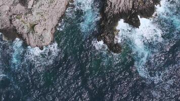 Aerial view of Capri's rugged coastline with cliffs, a natural arch, and vibrant blue waters. Waves crash dynamically near a structure with yellow rooftops. video