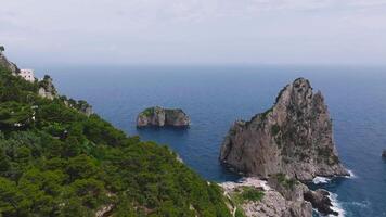 Smooth aerial motion over turquoise waters reveals the Faraglioni rocks, rugged cliffs, lush vegetation, and a small structure near the cliffs. video