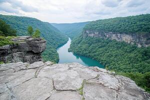 High viewpoint over a serene river valley. Lush green hills surround a winding, deep blue river, nestled within a dramatic canyon. Rocky outcrop offers a panoramic vista photo