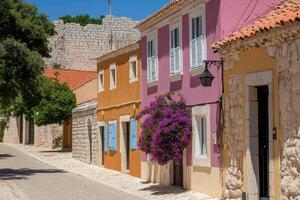 Colorful houses line a sunny street, shaded by trees, with a glimpse of a historic stone wall in the background photo