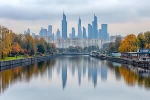 Autumn cityscape reflected in a calm river. Modern buildings mirrored in tranquil water, flanked by colorful fall foliage photo