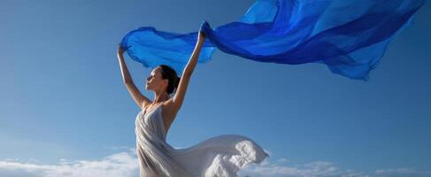 The graceful woman dancing with a flowing blue scarf against a bright sky. photo