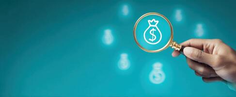 The hand holding a magnifying glass over money symbols on a turquoise background. photo
