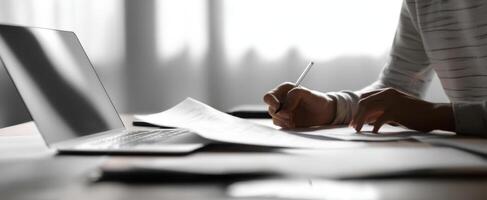 The person writing notes beside a laptop and scattered documents on the desk. photo