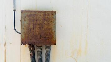 Rusty square metal box mounted on a weathered white wall. Power and signal cables are connected to a connection set inside a metal box. Covered with a black plastic flex tube. Install on cement wall. photo