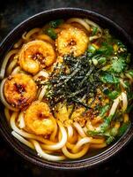 Delicious Udon Noodle Soup with Tempura Shrimp Close Up. A top view image showing a bowl of udon noodles in a rich broth, garnished with tempura shrimp, seaweed, and cilantro. photo
