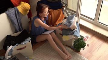 A woman sits on the floor surrounded by bins clothes and checklists thoughtfully analyzing each item while building a mindful wardrobe using slow fashion habits simplicity and intentional decisions video