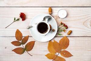 A flat lay image featuring a cup of black coffee on a saucer, surrounded by autumn leaves, acorns, a red flower, and a small candle on a light wooden surface. photo