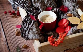 A close-up, overhead shot of a wooden box filled with a textured grey scarf, a red-handled mug of coffee, red macarons, rowan berries, a yellow leaf, and an acorn. photo