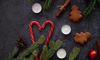 A festive flat lay featuring Christmas decorations, including candy canes forming a heart shape, gingerbread tree cookies, cinnamon sticks, pine branches, candles, and a red ornament photo