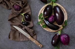 A photograph of a culinary setting with eggplants and parsley, with a knife, set against a textured, grey background. photo