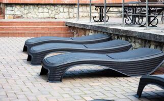 The image shows a row of modern black wicker lounge chairs neatly arranged on a paved patio area next to a stone and brick wall. photo