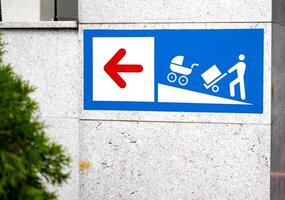 Sign with a red left arrow and icons showing a person with a cart and stroller on a ramp. Placed on a stone wall background. Accessibility concept photo