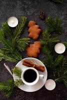 A photograph of a Christmas setting with a coffee cup, gingerbread cookies, cinnamon sticks, candy canes, candles, and pine sprigs set against a dark, textured surface. photo