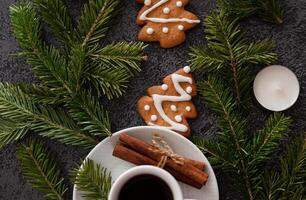 A simple yet elegant flat lay depicting Christmas decorations a cup of coffee, gingerbread cookies, cinnamon, candy canes, candles, and pine branches neatly arranged on a dark background. photo