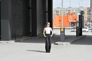 A solitary figure in fashionable black wide-leg pants and a white sleeveless top, standing on an empty paved area. photo