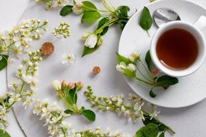 The image depicts a delicate arrangement of small white blossoms and green leaves scattered elegantly on a white surface, surrounding a white cup filled with tea placed on a matching saucer. photo